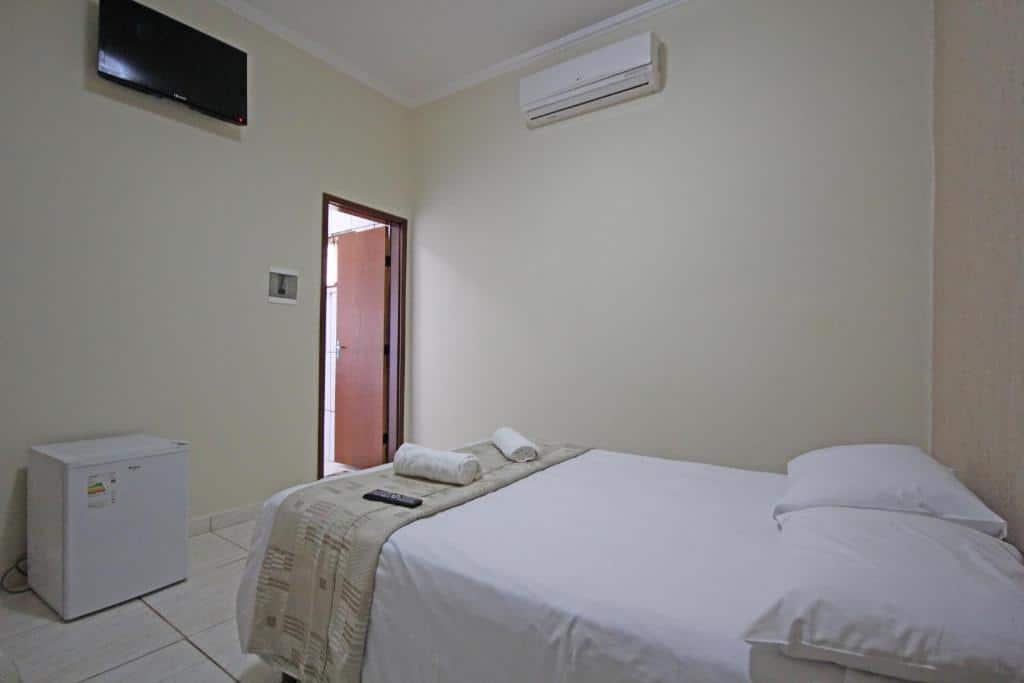 Um pequeno quarto da Pousada Facincani, uma das pousadas em Olímpia, com uma cama, frigobar, TV na parede, ar-condicionado e uma porta aberta para o corredor.