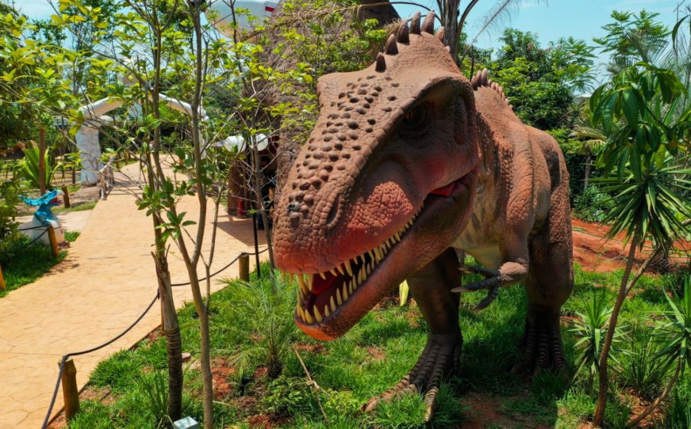 Uma estátua de dinossauro grande e realista está entre plantas e árvores verdes ao ar livre em um dia ensolarado no Vale dos Dinossauros em Olímpia.