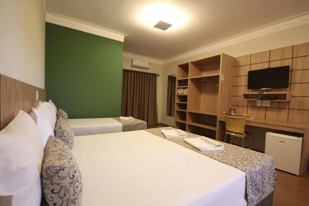 Quarto da Pousada Vitalis, um das pousadas em Olímpia, com duas camas, parede com detalhes verdes, escrivaninha, TV, frigobar e prateleiras, iluminado pela luz do teto.