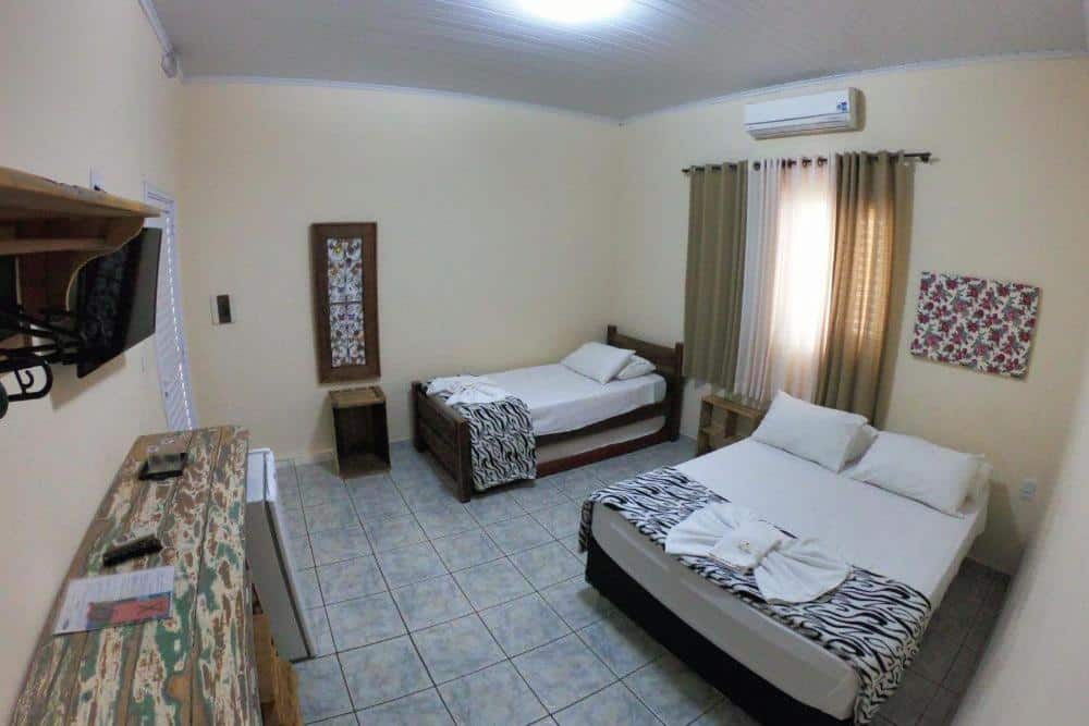 Quarto da Pousada Tia Nena, uma das pousadas em Olímpia, com uma cama de casal e uma cama de solteiro, TV, cômoda, duas janelas com cortinas e piso de azulejo.
