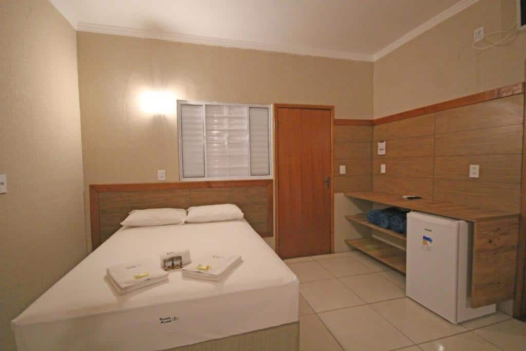 Um quarto da Pousada Recanto da Morada, uma das pousadas em Olímpia, com uma cama de casal, toalhas dobradas, uma porta de madeira, prateleiras e uma pequena geladeira branca.