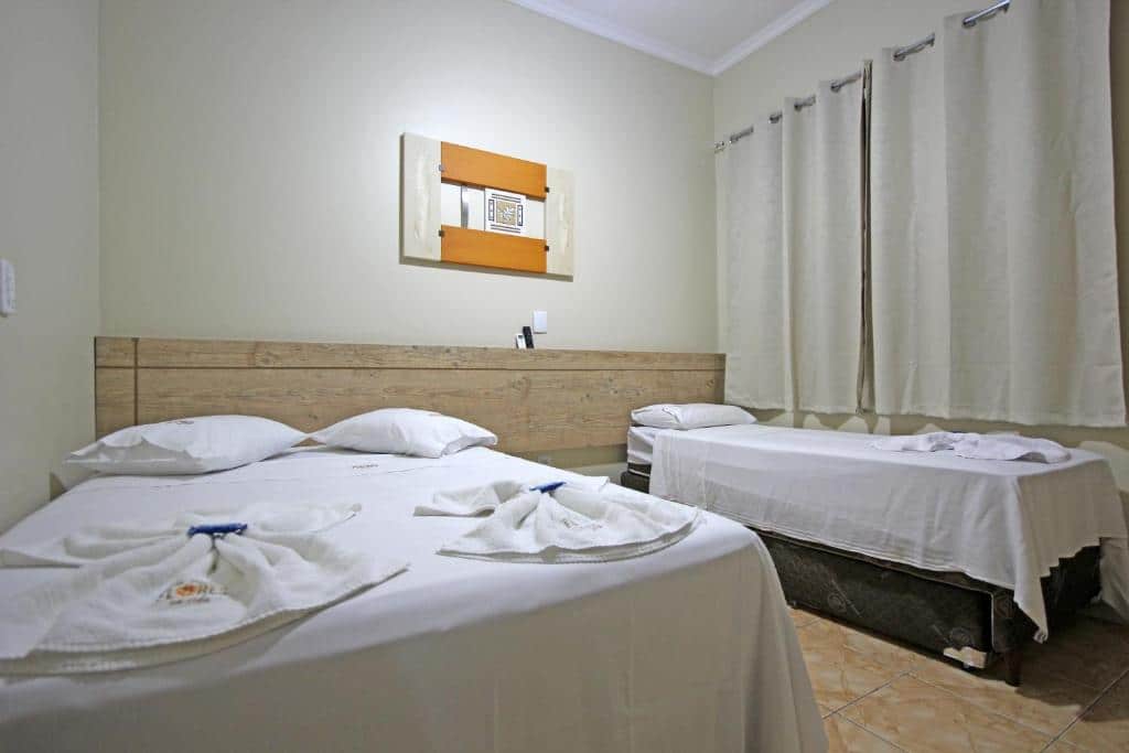 Um quarto da Pousada Flores em Vida, uma das pousadas em Olímpia, com uma cama de casal e uma cama de solteiro, ambas cobertas com lençóis e toalhas brancos e cortinas bege na janela.
