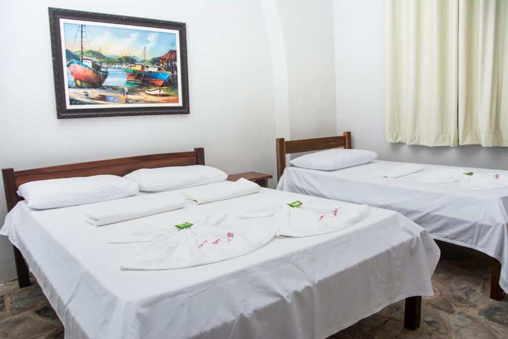 Quartos da Pousada Porto das Barcas, em Parnaíba, com duas camas bem arrumadas com lençóis brancos em um quarto simples, um quadro e cortinas creme na parede.