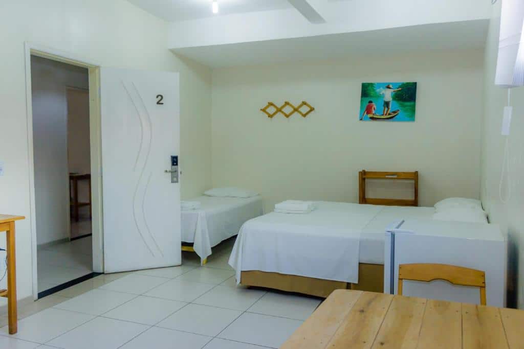 Quarto da pousada Vila do Carmo, em Parnaíba, sendo ele simples com duas camas, uma pequena mesa, piso de azulejos brancos, porta aberta e arte na parede acima das camas.