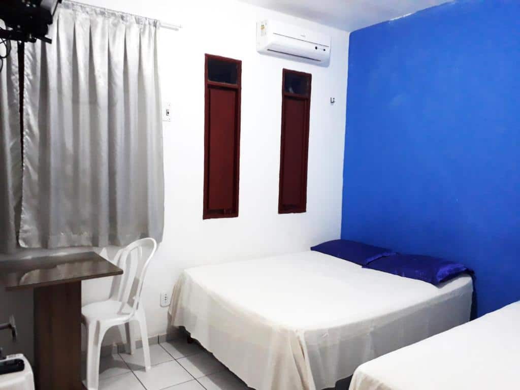 Quarto da Pousada Lunar com duas camas, uma pequena mesa com uma cadeira de plástico branca, parede com detalhes em azul, cortinas prateadas e ar-condicionado.