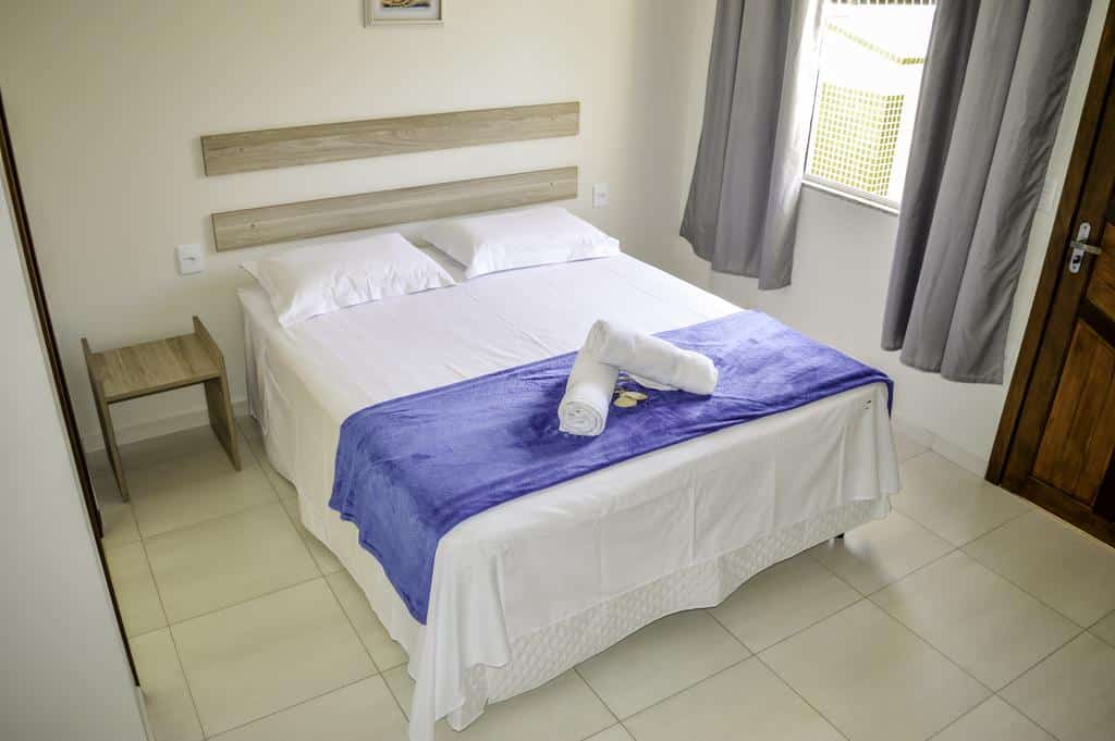 Quarto da Pousada Vila Regina com cama bem arrumada com lençóis brancos, um cobertor roxo e toalhas, ao lado de uma janela e porta de madeira em um quarto simples. Ilustra o post de pousadas em Penha.