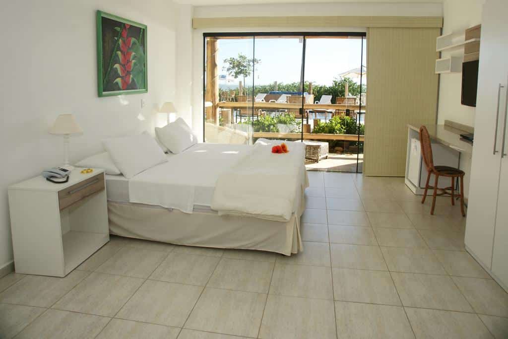 Um quarto da Pousada Pedra da Ilha arrumado com uma cama de casal, escrivaninha, cadeira e grandes portas de vidro que se abrem para um pátio com assentos ao ar livre. Ilustra o post de pousadas em Penha.