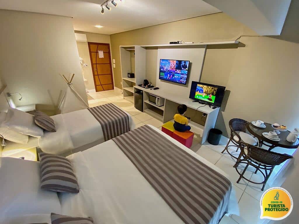 Quarto da Pousada Molinha com duas camas, uma criança assistindo à TV, uma pequena área de refeições e um banheiro visível ao fundo. Ilustra o post de pousadas em Penha.