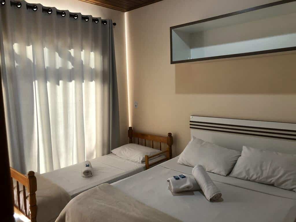 Um pequeno quarto da Pousada Rosália com uma cama de solteiro e uma cama de casal, ambas com lençóis e toalhas brancos, perto de uma janela com cortinas fechadas. Ilustra o post de pousadas em Penha.