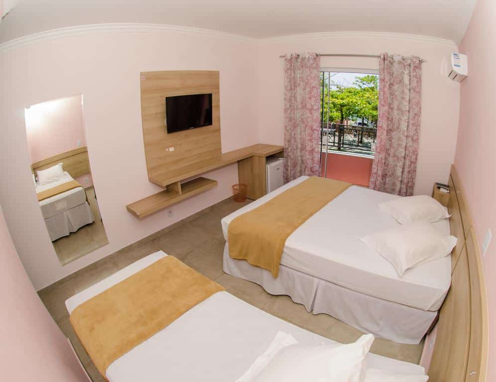 Quarto da Pousada Baía dos Açores com uma cama de casal, uma cama de solteiro, uma TV na parede, um espelho e uma janela com cortinas florais. Ilustra o post de pousadas em Penha.