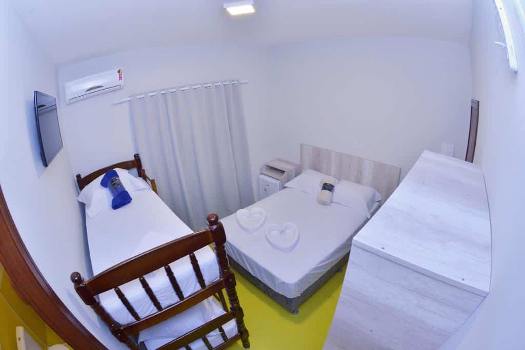 Quarto da Pousada Recanto das Cores com uma cama de solteiro, uma cama de casal, móveis brancos, TV e toalhas dobradas em forma de coração sobre as camas.