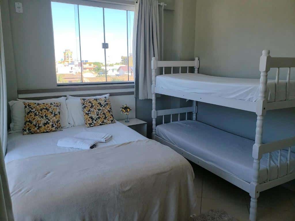 Quarto da Pousada Recanto do Cowboy com uma cama de casal, dois travesseiros florais, beliche branco, mesa de cabeceira e janela com vista para a cidade. Ilustra o post de pousadas em Penha.