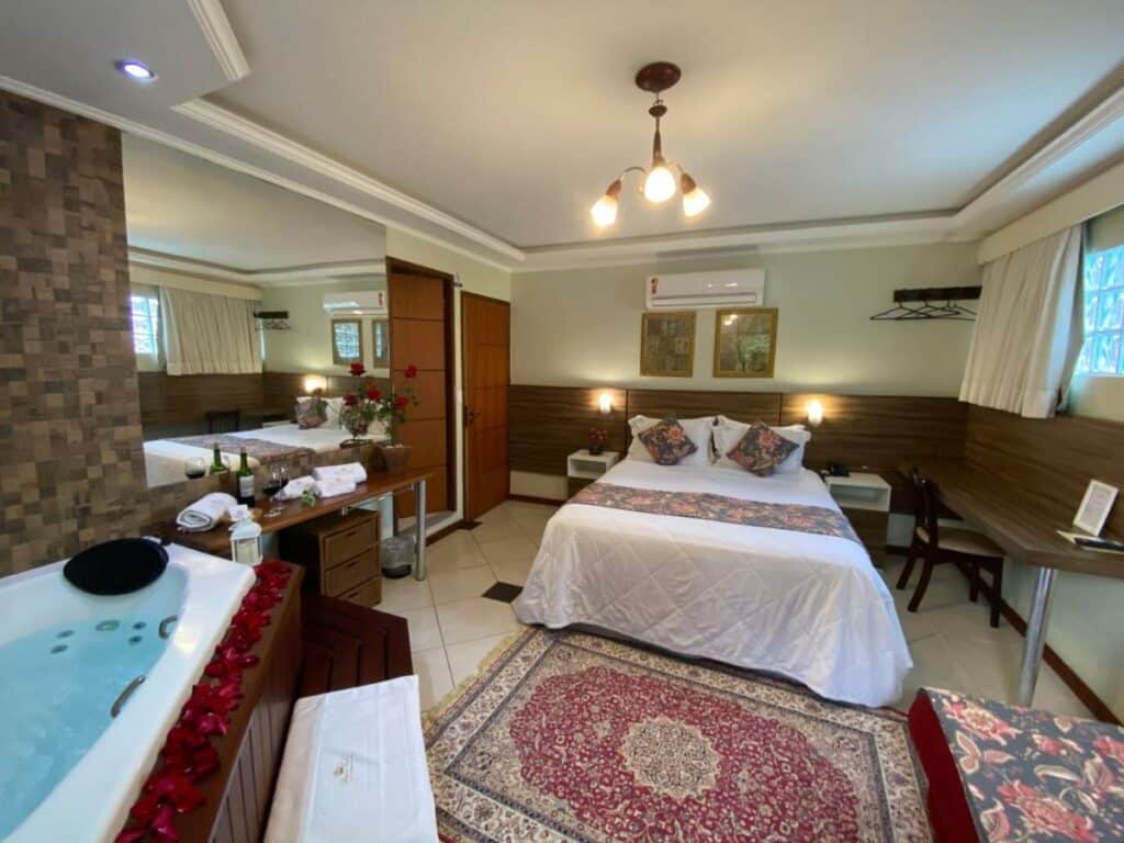 Quarto Pousada Pousada Princesa Isabel Dom Pedro, com cama de casal com leçóis brancos e travesseiros coloridos. Quadros na acima da cabeceira na parede e um ar-condicionado, uma banheira no canto do quarto perto de algumas pétalas de rosa e de uma parede de pedras. Mesa de cabeceira com abajour, estante com toalhas e decoração.
