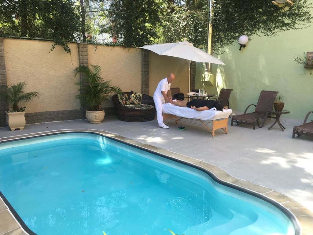Piscina Pousada Princesa Isabel, área da piscina, com duas espreguiçadeiras em segundo plano, a piscina ao centro, ao fundo um hóspede deitado em uma maca recebe uma massagem de um funcionário de branco.