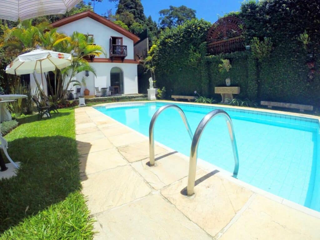 Área da piscina da Pousada Koeler, piscina com água azul cercada por um gramado e ao fundo a parte de trás da pousada. 