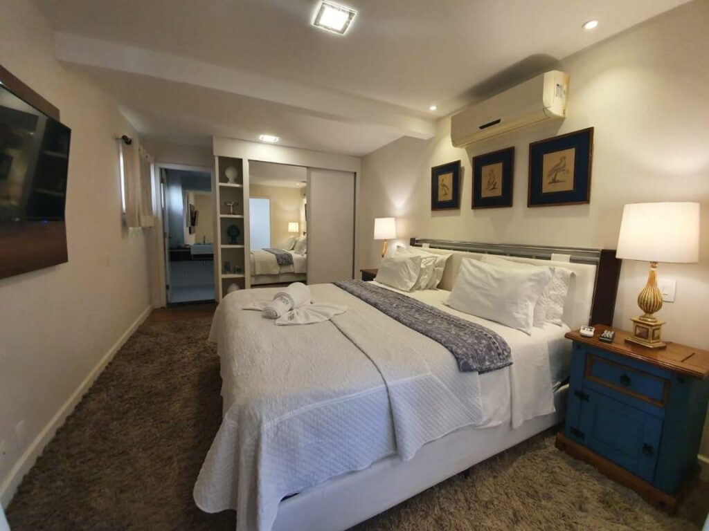 Quarto da Pousada Orquídea da Serra, cama king size com lençóis brancos e cinza, com quadros na parede e também um ar-condicionado, mesa de cabeceira com abajour e entrada do banheiro no canto. 