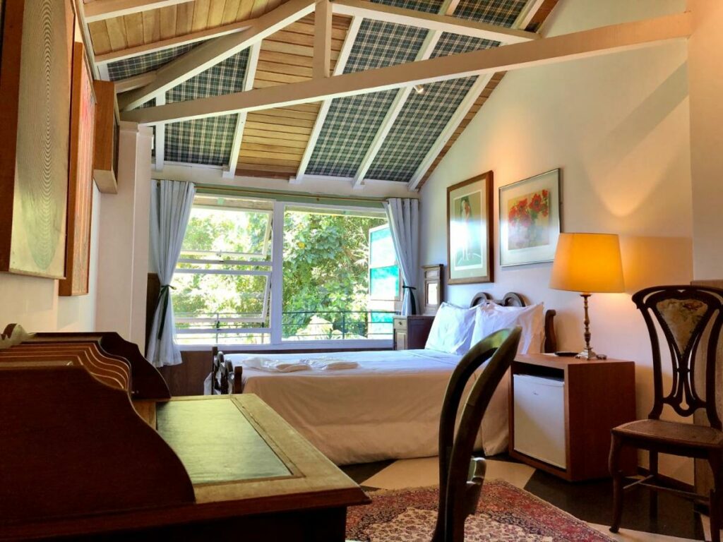 Quarto da Pousada Pataca de Prata, com cama ao centro com lençóis brancos, mesa de cabeceira e janela ao canto. 