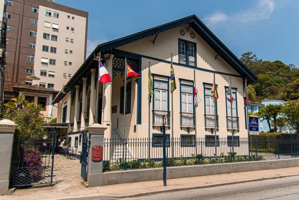 Faixada da Pousada Dom Petrópolis, aparenta ser uma casa antiga, com escadaria, janelas grandes, várias bandeiras a frente decorando a faixada. 