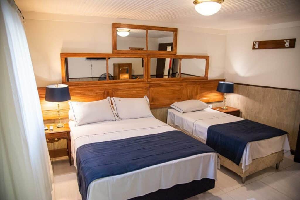 Quarto com cama de casal e uma de solteiro na Pousada Dom Petrópolis, com espelhos na cabeceira de madeira, mesa de canto, com abajour e cortina ao canto. 