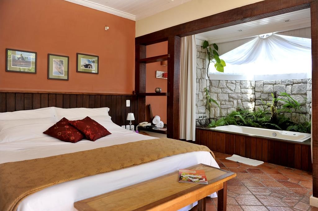 Quarto Pousada Recanto das Araras, com cama de casal com leçóis brancos e travesseiros vermelhos. Quadros na acima da cabeceira na parede, uma banheira no canto do quarto perto de algumas plantas ornamentais e de uma parede de pedras. Mesa de cabeceira com abajour, estante com toalhas e decoração.