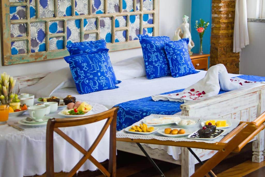 Uma cama da Pousada Solar dos Deuses com lençóis azuis brilhantes, bandejas de café da manhã com alimentos e uma toalha dobrada em forma de cisne estão em um quarto aconchegante e decorado.