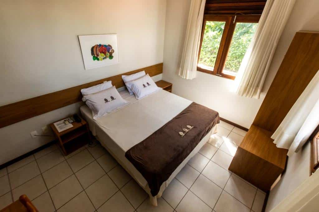 Uma cama de casal na Pousada Pedra da Concha bem arrumada com dois travesseiros, cobertor marrom, mesinhas de cabeceira e uma janela que permite a entrada de luz natural.