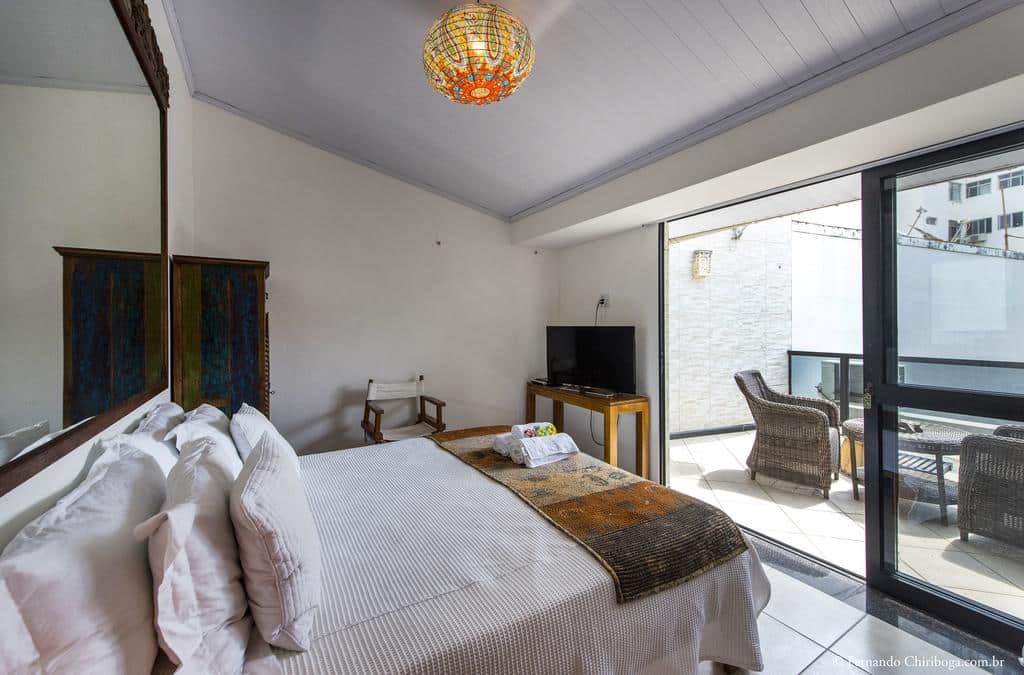 Um quarto da Casa Petunia Pousada Boutique com cama, TV, cadeira e porta do pátio que leva a uma área de estar externa com móveis de vime.