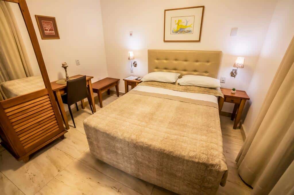 Uma cama bem arrumada em um pequeno quarto da Pousada Bahia Pelô com duas mesinhas de cabeceira, uma escrivaninha e quadros de arte na parede.