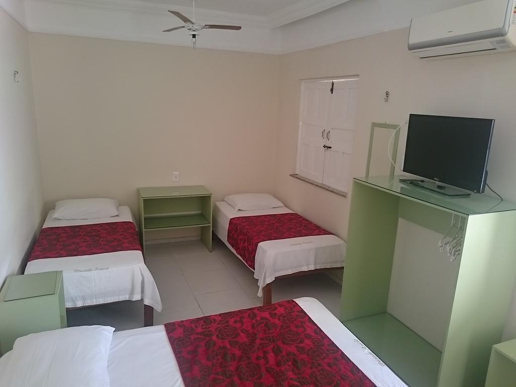 Um quarto simples da Pousada Acácia da Barra com três camas de solteiro, uma pequena estante verde, uma TV em um suporte e um ar-condicionado na parede.