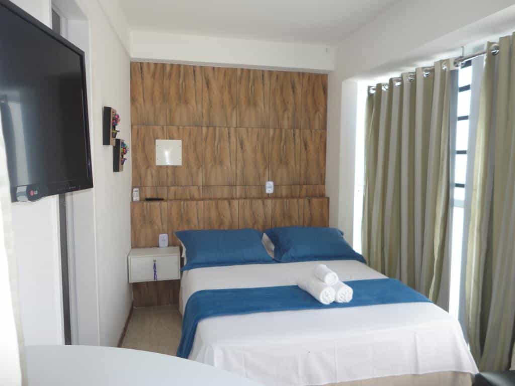 Quarto da Pousada Pedacinho da Bahia moderno com uma cama de casal, roupa de cama azul, uma TV na parede e janelas grandes com cortinas bege.