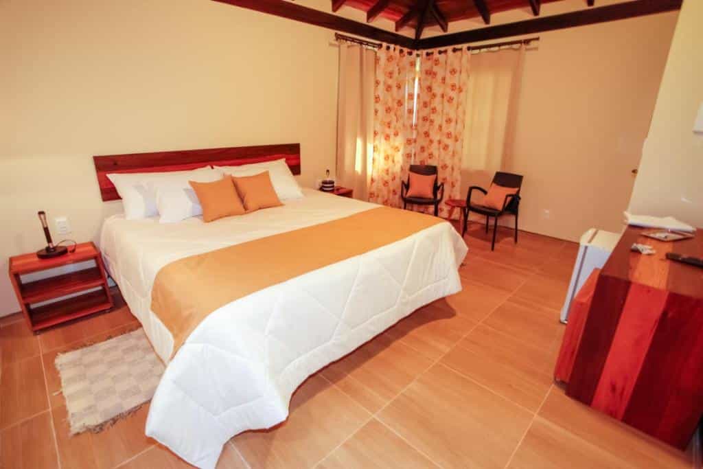 Um quarto na Villa Severina (Brumadinho) com uma cama grande, duas cadeiras ao lado de uma janela com cortinas florais, uma escrivaninha de madeira e uma mesa de cabeceira.