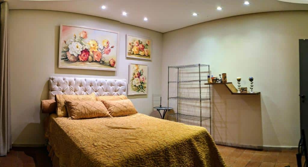 Um quarto no Sítio Esperança e Fé (Ribeirão das Neves) com uma cama, colcha dourada, pinturas florais, uma prateleira de arame e uma prateleira montada na parede com itens decorativos.