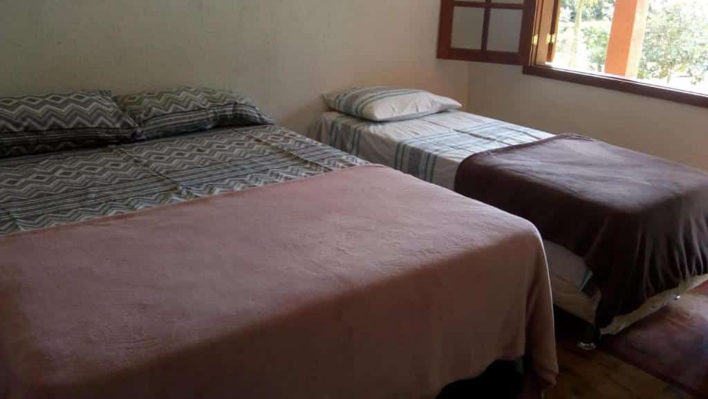 Duas camas com cobertores e travesseiros em um quarto na Pousada Fazenda Bocaina (Inhaúma) com uma janela aberta e luz vinda de fora.