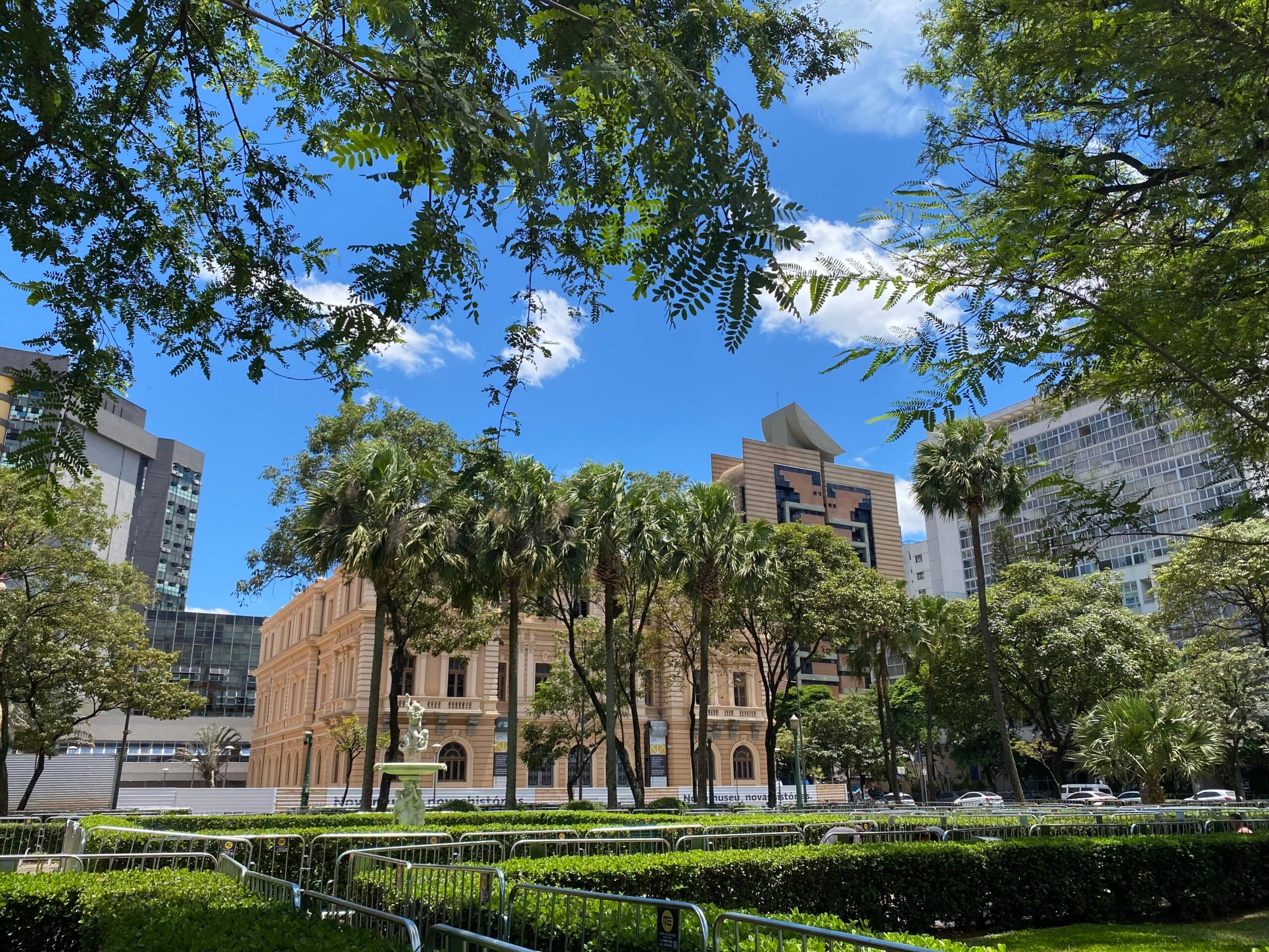 Edifício histórico com janelas em arco cercado por árvores sob um céu azul, com edifícios modernos ao fundo.
