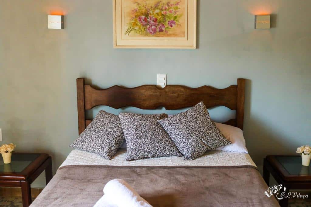 Uma cama bem arrumada com quatro travesseiros estampados, cobertor marrom e uma toalha, ladeada por duas mesas laterais, sob uma pintura floral, em um dos quartos da Pousada e Restaurante Victórios (Caeté).