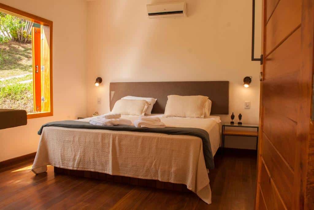 Uma cama bem arrumada em um quarto iluminado pelo sol, com piso de madeira, dois travesseiros e uma janela aberta mostrando a vegetação do lado de fora, em um dos quartos da Villa Domaso (Brumadinho), para representar pousadas perto de BH.
