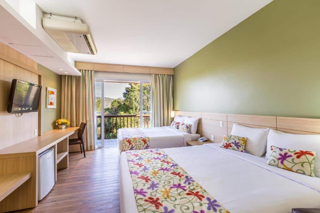 Quarto do Tauá Hotel & Convention Caeté (Caeté) com duas camas, escrivaninha, TV na parede, frigobar, varanda e parede com detalhes em verde, para representar pousadas perto de BH.