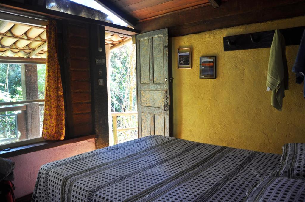 Uma cama com um cobertor estampado em um quarto rústico com paredes amarelas, uma porta aberta e a luz do sol entrando pela janela, na Pousada Rural Lua Bonita do Nero (Macacos), para representar pousadas perto de BH.