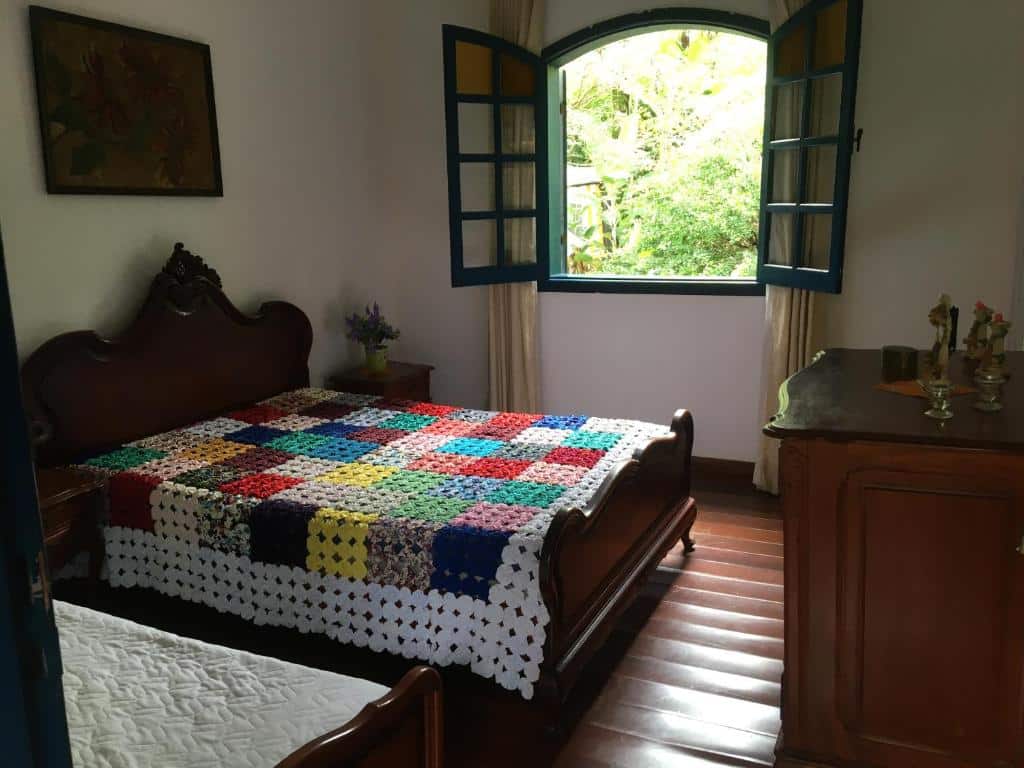 Quarto na Pousada Rancho Flores (Caeté) com uma cama de madeira, colcha de retalhos colorida, cômoda, flores e uma janela aberta mostrando a vegetação do lado de fora, para representar pousadas perto de BH.