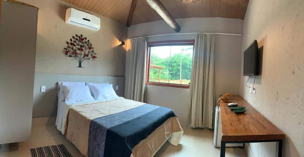 Uma cama bem arrumada em um quarto simples com janela, TV, escrivaninha, ar-condicionado e arte de parede de árvore acima da cama, em um dos quartos da Pousada Villa Recanto dos Lagos (Brumadinho), para representar pousadas perto de BH.
