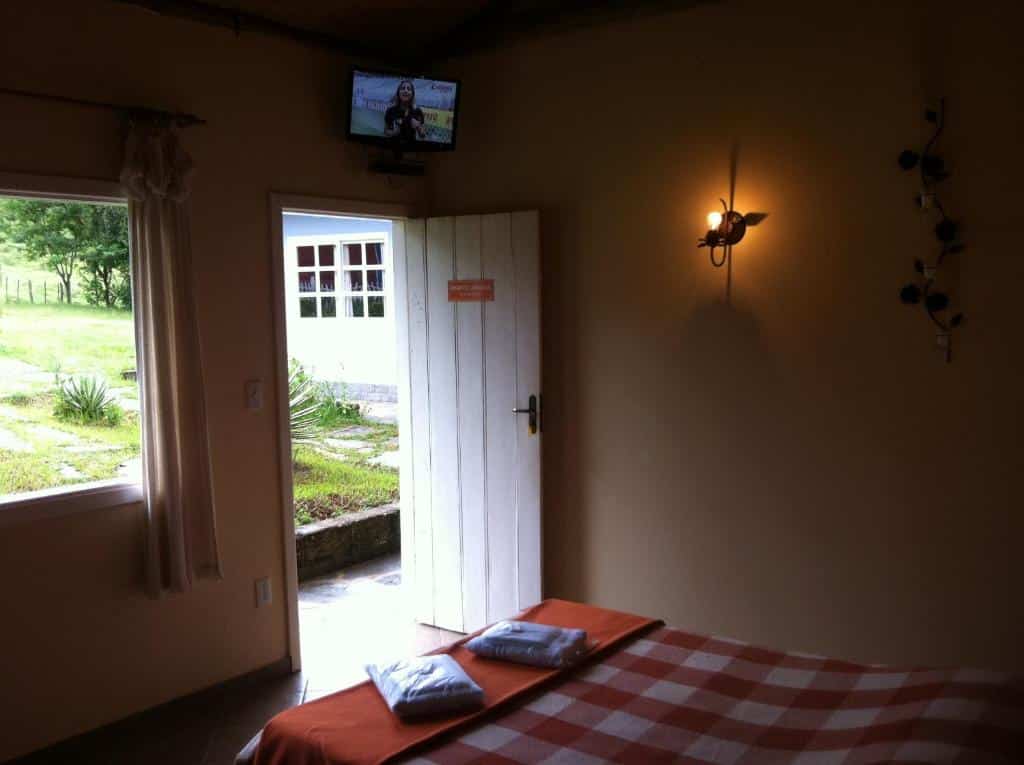 Um pequeno quarto na Pousada Tabuleiro Eco (Conceição do Mato Dentro) com uma cama, uma porta aberta para um jardim, uma TV na parede e uma lâmpada de parede acesa.