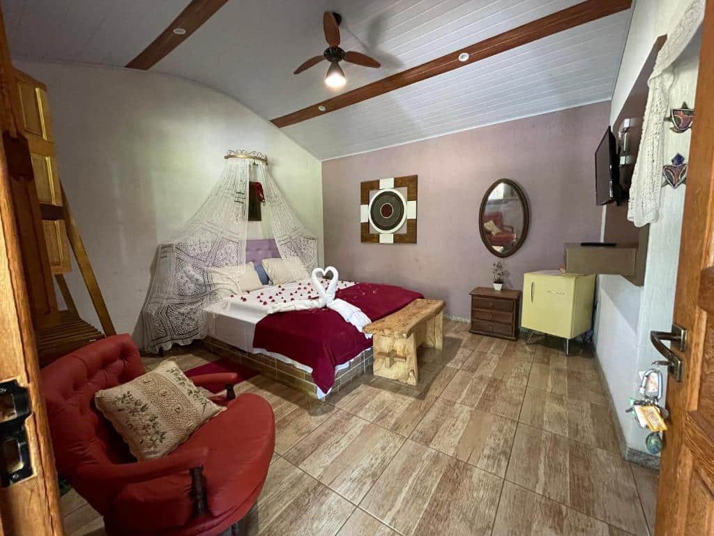 Um quarto aconchegante no Kafofinho da Serra (Serra do Cipó) com uma cama de casal, roupa de cama vermelha, mosquiteiro, poltrona, TV e móveis de madeira em um piso de azulejo, para representar pousadas perto de BH.