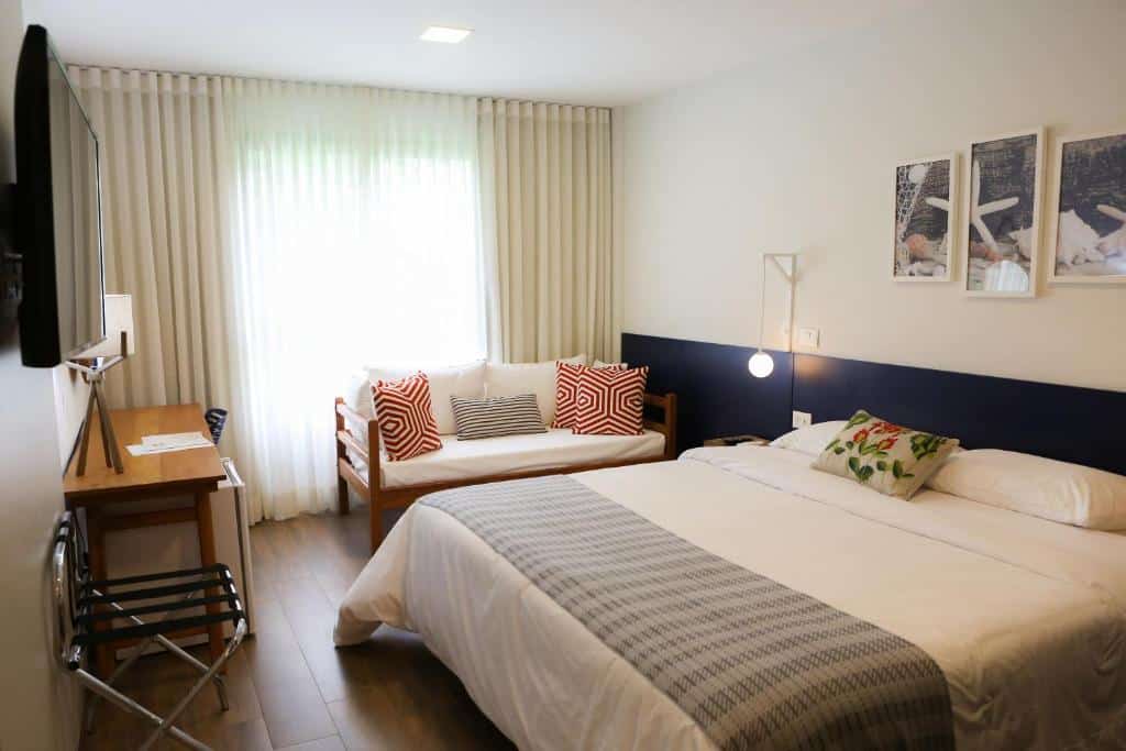Quarto do Hotel do Bosque ECO Resort com cama de casal, varanda com cortinas, um sofá com dois lugares com almofadas, uma televisão, uma mesa de trabalho com uma cadeira, um frigobar e alguns itens de decoração, para representar resorts em Angra dos Reis
