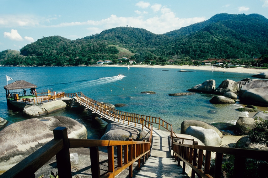 Descida que leva até um deck no Club Med Angra dos Reis, no local há saída de jet-ski e balsas, ao redor é possível ver algumas rochas e uma praia que, logo ao fundo, tem enormes montanhas verdes