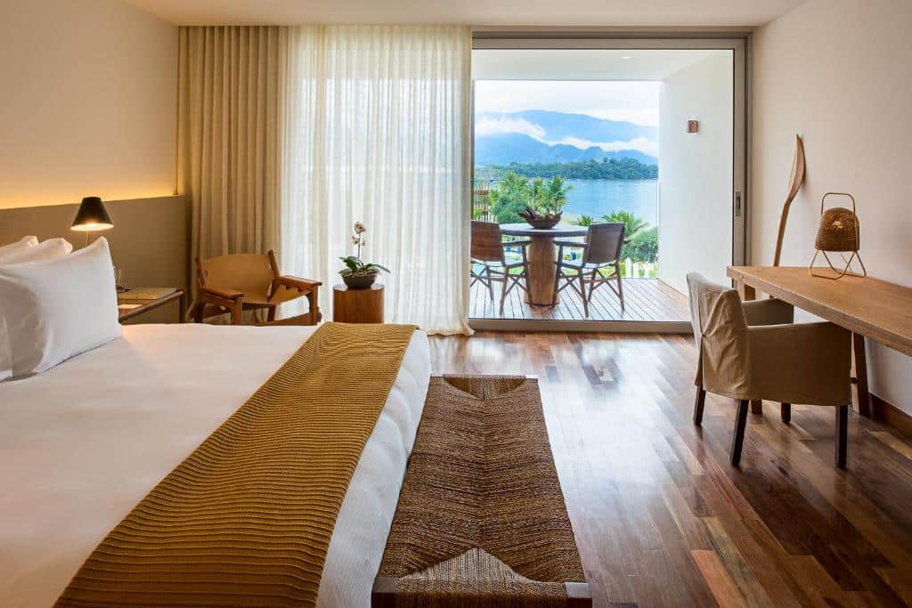 Quarto amplo do Hotel Fasano Angra dos Reis com piso em madeira, varanda com vista para o mar e montanhas com uma mesa com duas cadeiras, dentro há um poltrona e uma bancada de madeira, uma cama de casal e poucos itens de decoração rústicos e em madeira