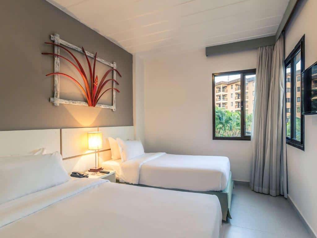 Quarto do Mercure Angra dos Reis com duas camas de solteiro, piso frio, uma janela de canto que oferece uma boa vista, uma televisão na parede de frente com as camas