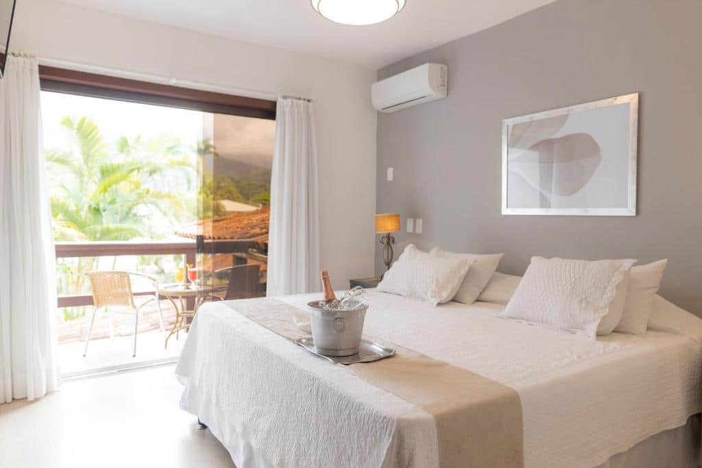 Quarto do Angra Boutique Hotel com uma varanda com porta de vidro, com uma mesinha e duas cadeiras, do lado de dentro há uma cama de casal, uma mesinha de cabeceira com um abajur e um ar-condicionado de teto