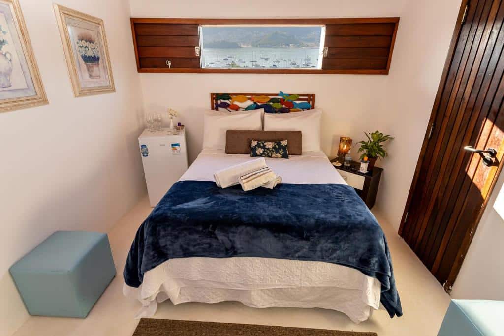 Quarto da Rede Reserva Costa Verde com uma cama de casal com almofadas e toalhas sob ela, um frigobar do lado esquerdo, um mesinha com abajur do lado direito, há alguns itens de decoração e dois bufês para se sentar