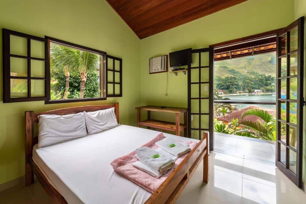 Quarto do Praia e Mar Angra  com uma varanda ampla dando vista para o mar, uma cama de casal com toalhas e travesseiros, um pequeno móvel de madeira com prateleiras do lado direito, além de uma televisão e um ar-condicionado de teto