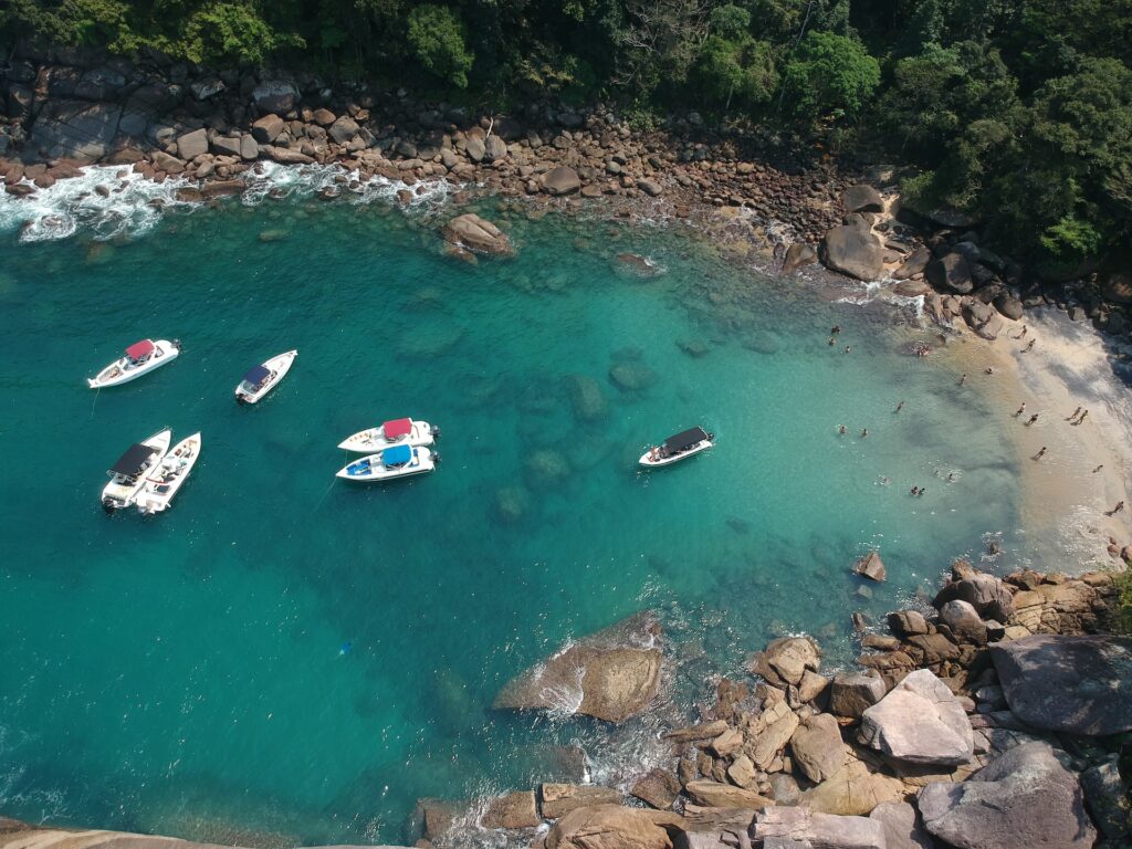 Vista aérea da praia do Cachadaço em Angra dos Reis, é um local com uma faixa de areia bem pequena e com muitas pedras e montanhas ao redor, a água é transparente e há algumas balsas paradas